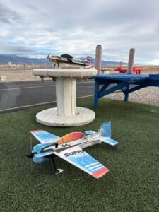 Two RC foam 3D aerobatic airplanes, including a Supra Race model, sitting at an outdoor flying field with benches and mountains in the background.