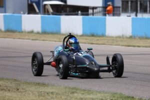 Open-wheel Vortech Formula Vee race car on track with a helmeted driver taking a corner during a road racing event.